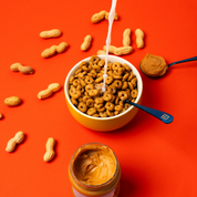 Bol de cereales Magic Morning saveur peanut butter avec du lait verse, accompagne de cacahuetes entieres, d’une cuillere de beurre de cacahuete et d’un pot ouvert sur fond orange. Une option saine et proteinee pour un petit-dejeuner faible en sucre.